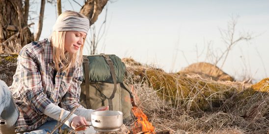 Hiking woman with backpack cook by campfire Young hiking woman with backpack cook on campfire in countryside